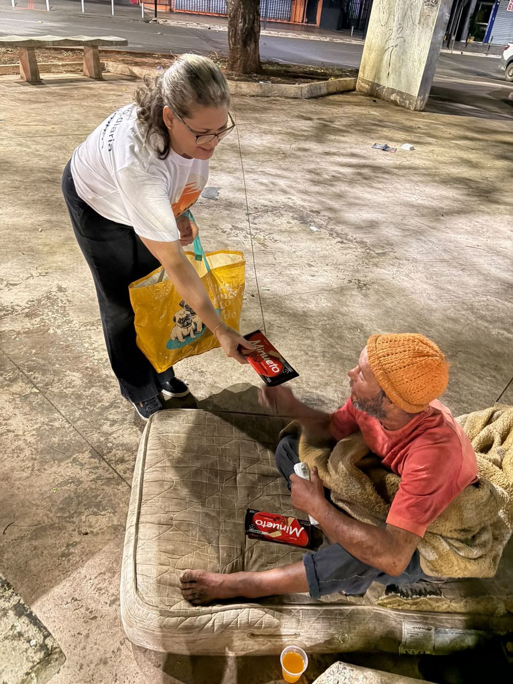 Voluntária entregando chocolate para um senhor sentado num colchão na praça