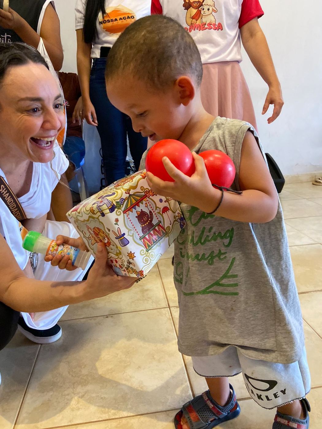 Voluntária sorrindo enquanto um menino abre o presente com bolas vermelhas